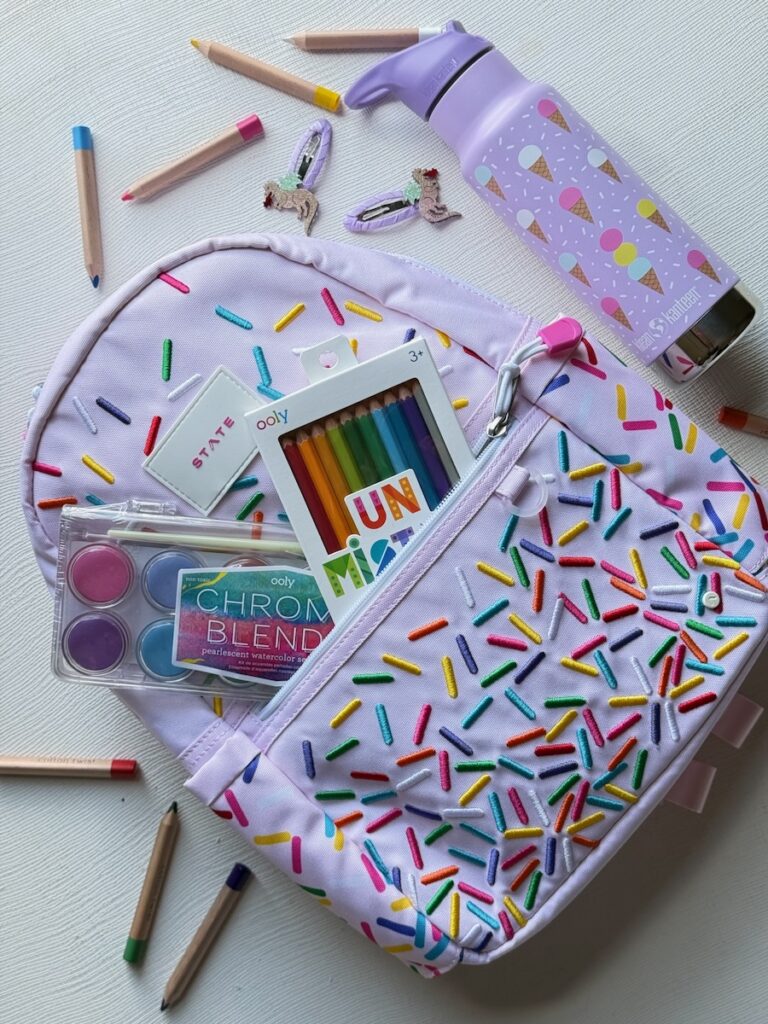 A purple backpack with rainbow sprinkles all over it and craft supplies in the pocket with a purple water bottle beside the backpack.