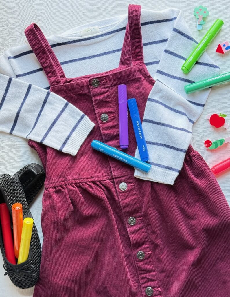 A kids red tank top dress over a striped long sleeve shirt with markets around the dress and in the dress shoes.