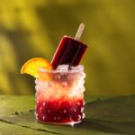 A short glass with a dark red blackberry bourbon bramble cocktail with a blackberry popsicle sticking out of the glass and an orange slice on the rim.