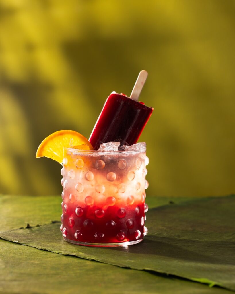 A short glass with a dark red blackberry bourbon bramble cocktail with a blackberry popsicle sticking out of the glass and an orange slice on the rim.