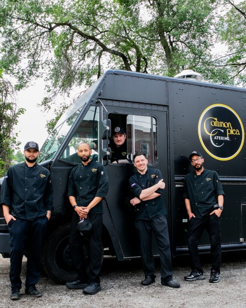 The Common Plea Catering culinary team stands with four members in black outside the catering truck and one member in the drivers seat of the truck.