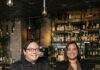 Cork & Crust at The Oaklander Excels in Food, Wine, and Cocktails: Head of the Table, July 2026 Kristen Butterworth and Amnelis Cruz sit side by side at a table in Cork and Crust with wine racks behind them.
