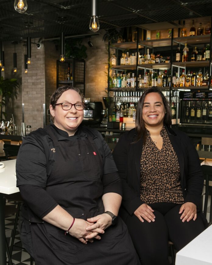 Cork & Crust at The Oaklander Excels in Food, Wine, and Cocktails: Head of the Table, July 2026 Kristen Butterworth and Amnelis Cruz sit side by side at a table in Cork and Crust with wine racks behind them.