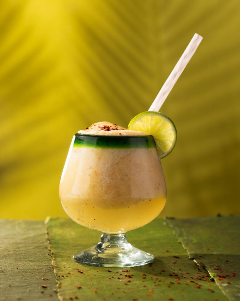 A short cocktail glass with a frozen Dole-Whip cocktail with a lime garnish and straw on a green background.
