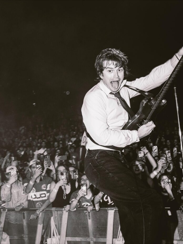 Djo plays guitar at a concert in a white shirt and black pants with his tongue out and a crowd behind him.