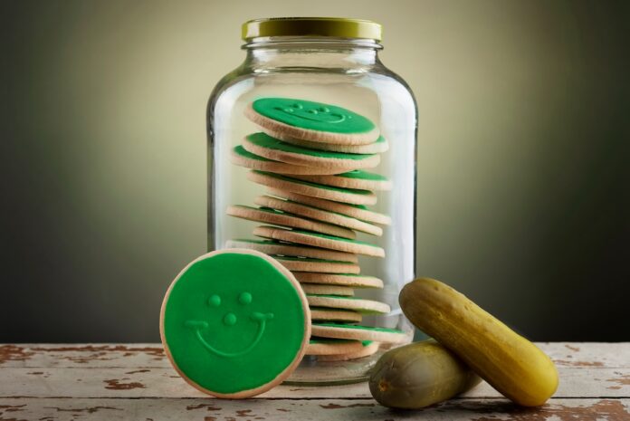 A pickle jar full fo pickle green smiley cookies on a table and green background with two pickles beside it.