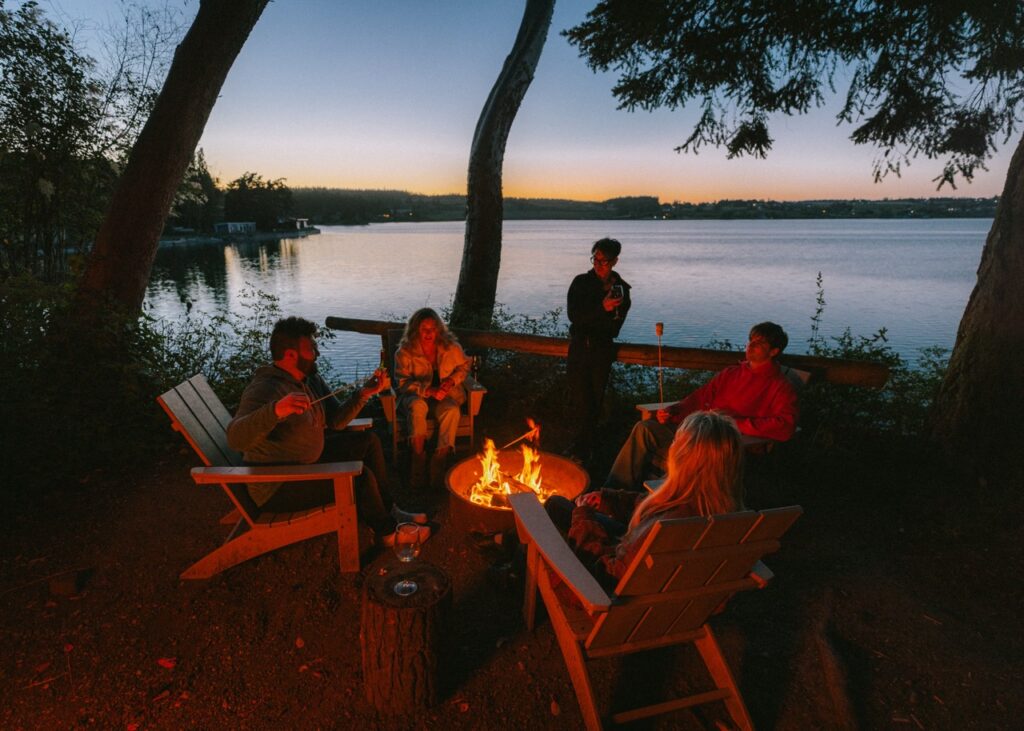 A group of people sit around a campfire by the lake.