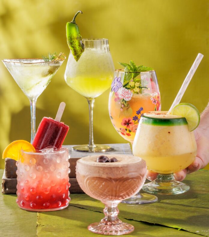 Frozen Cocktail Recipes for a Sweltering Pittsburgh Summer TABLE Magazine A set of six frozen cocktails in various glasses with summery toppings all on a green background.