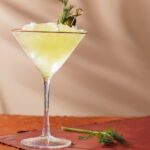 A tall martini glass with a frozen dirty martini, a gherkin garnish, and sprig of dill both inside the glass and on the table.