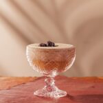 A short cocktail glass with a frozen Espresso Martini with three coffee beans on top on a pink background.