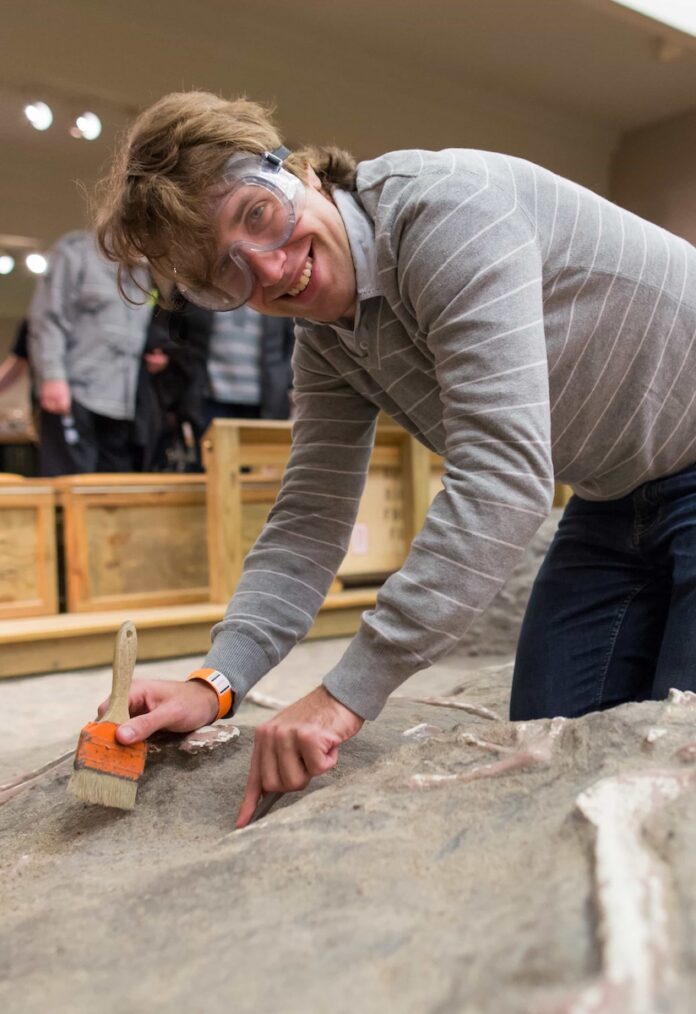 A man with goggles and a paint brush searches through sand for fossils at Camp Carnegie.