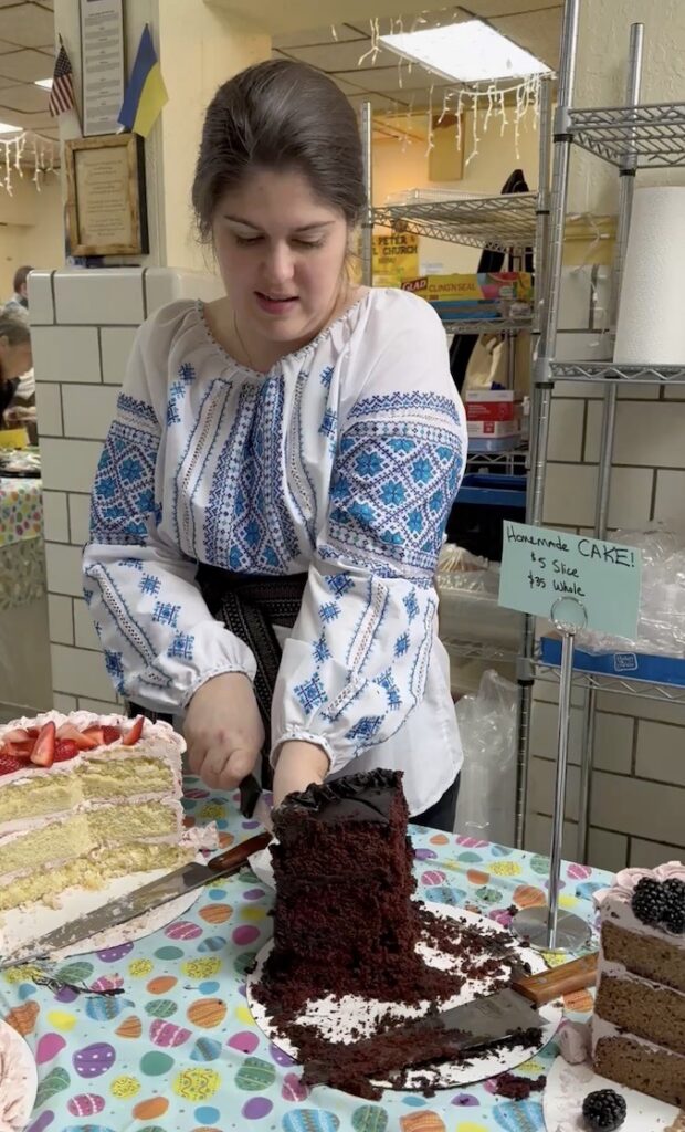 Woman in Ukrainian embroidered shirt cuts a chocolate cake at a Pittsburgh event.