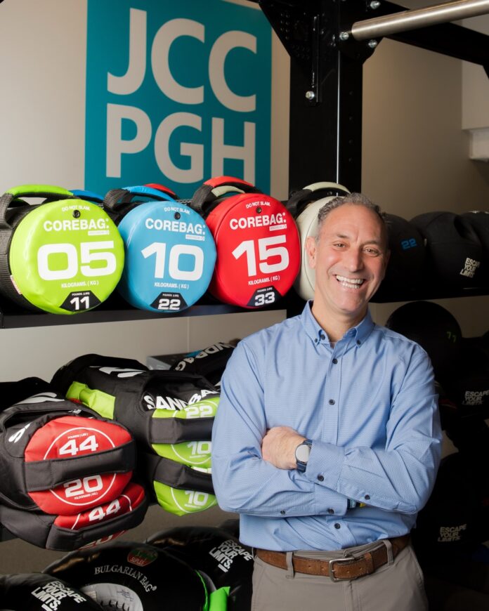 Jason Kunzman standing in front of the JCC PGH sign and bags of weights.