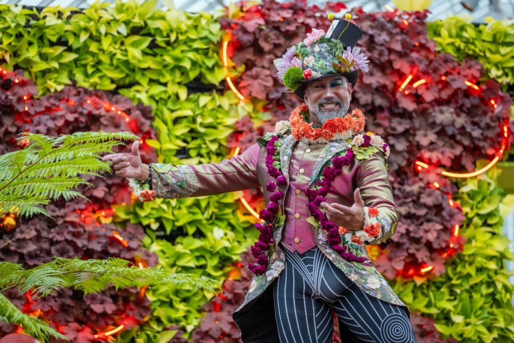 A man gestures dressed in a flower suit with a hat covered in flowers and greenery.