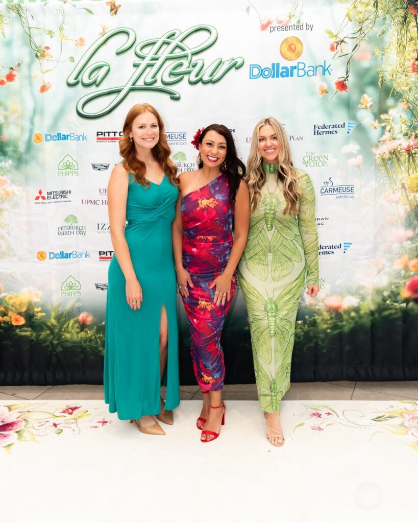 A woman in a blue dress with a woman in a floral dress and a woman in a green dress on a La Fleur backdrop.
