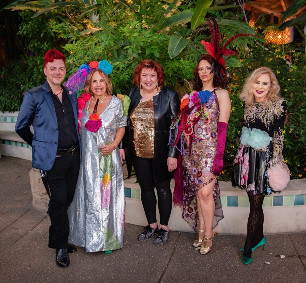 A group of four women and a man with red hair stand in front of greenery.