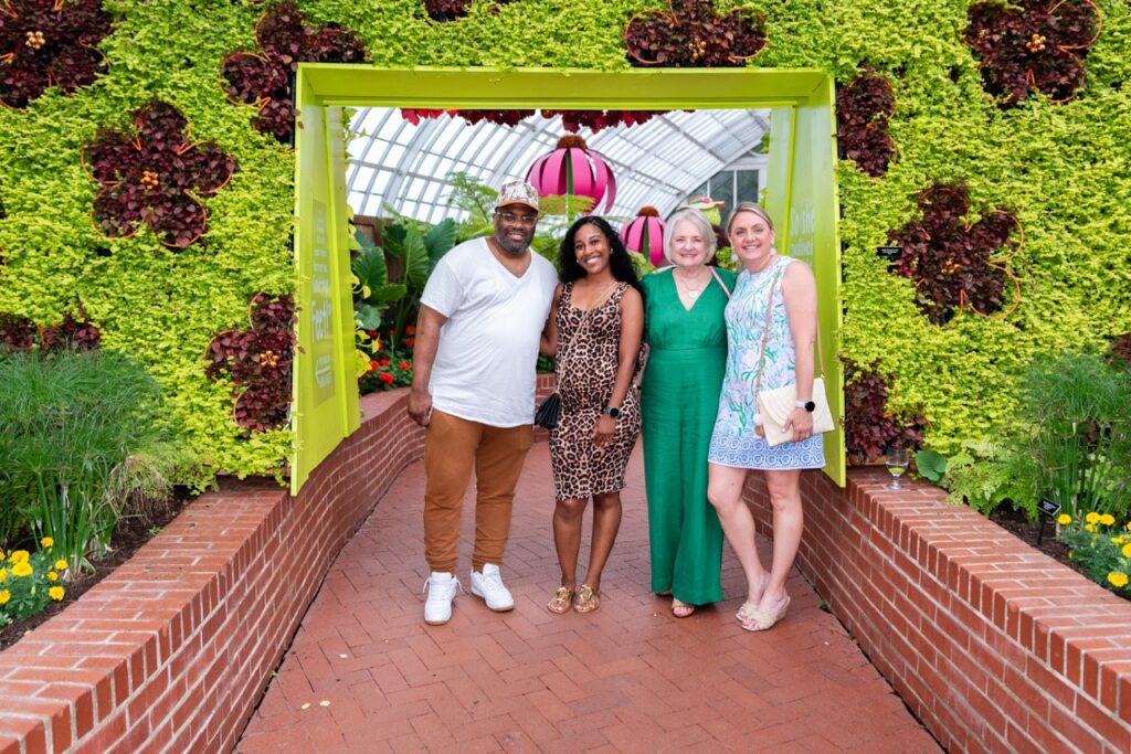 A couple in neutral color clothings stands with a woman in a green dress and a woman in a blue dress in front of a flower display.