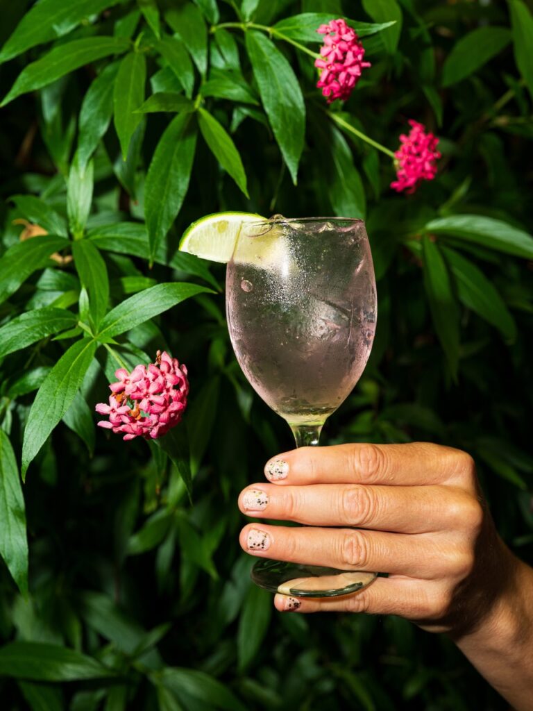 A woman holds a wine glass with a lime on the rim in front of a plant with pink blooms.