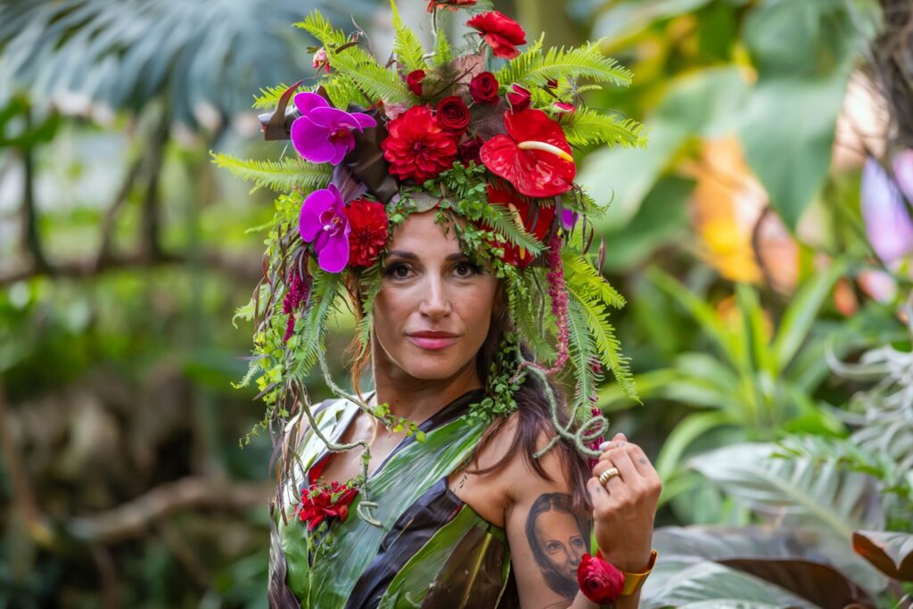 A man wears a flower and greenery head piece and leaves made into a top with a red flower on her wrist.