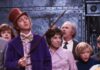 Movie Showings and Special Events Throughout Pittsburgh This July and August Willy Wonka stands beside a woman and children in the movie with his cane up in front of the woman who is wearing a jacket.