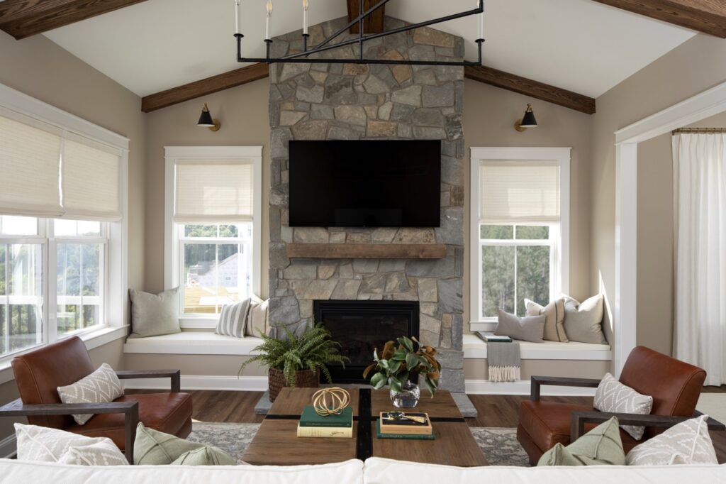A stone fireplace with a TV mounted on it in a room with many windows, two chairs, and a couch.