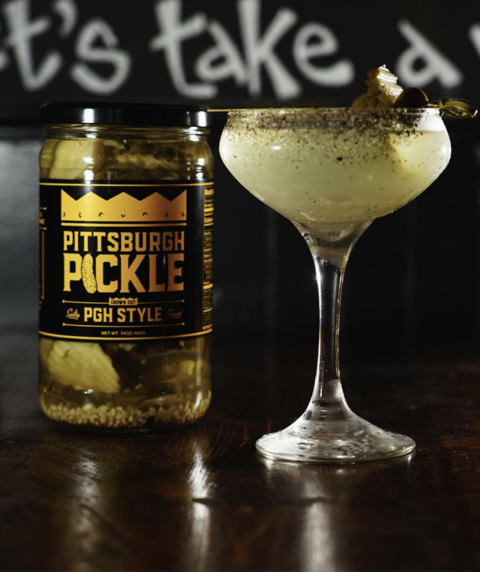 A pickle martini with a black salt rim on the glass beside a pickle jar from Pittsburgh Pickle Co.