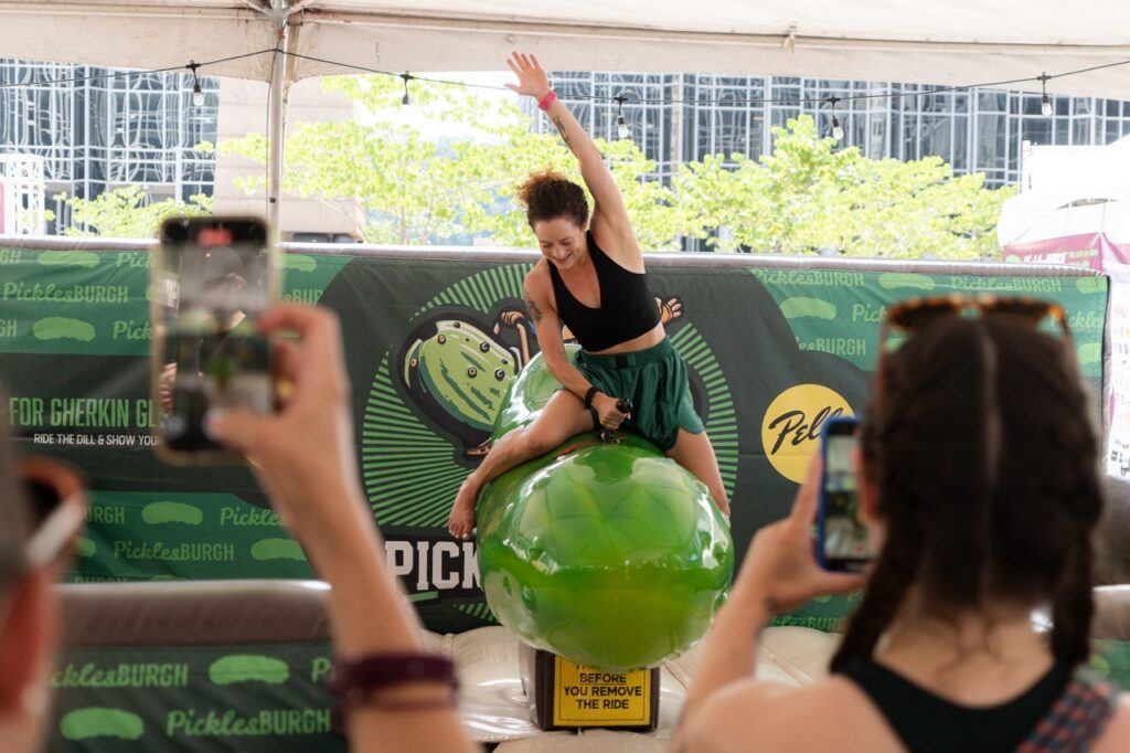 A woman rides a mechanical pickle in front of a Picklesburgh sign.