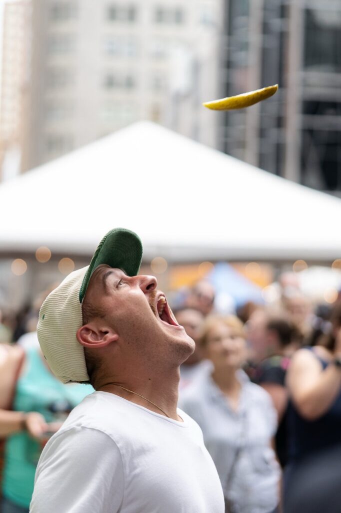 A man opens his mouth to catch a pickle spear flying through the air.
