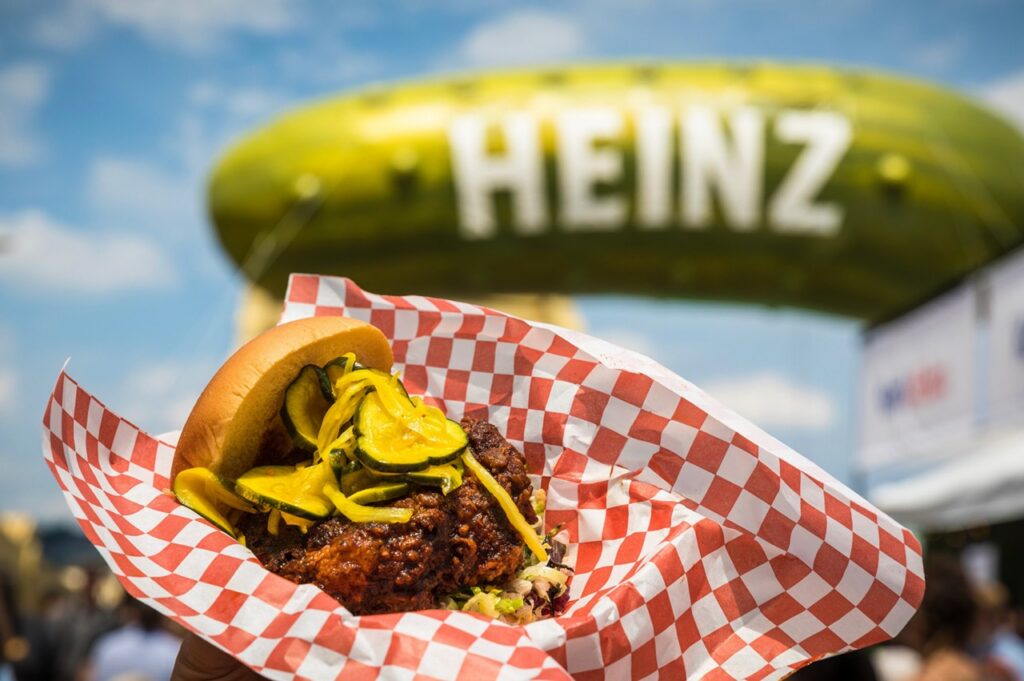 A chicken sandwich with pickles on it in front of the inflatable Heinz pickle balloon.