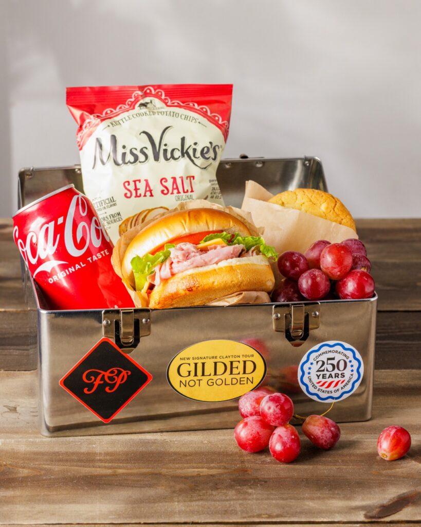 A steel lunch box with a sandwich, Coke, chips, and grapes inside.