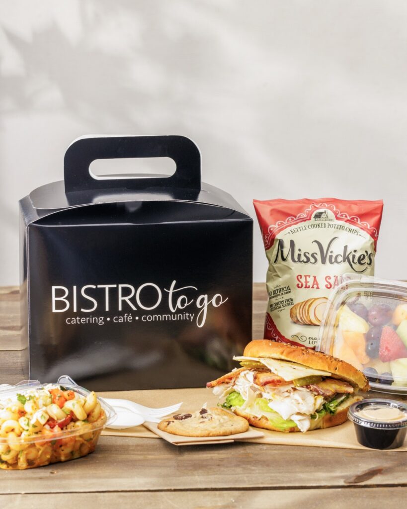 A black Bistro to Go box beside a bag of chips and a sandwich and cookie.