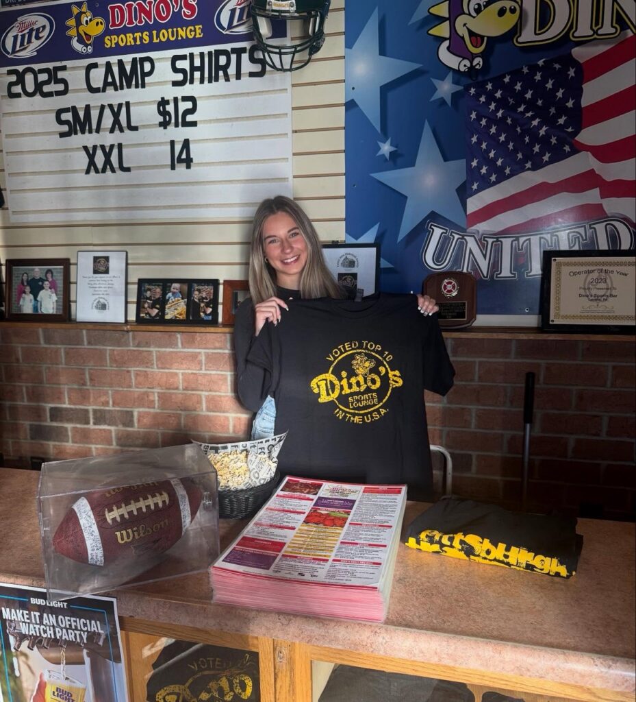 A woman holds up a Dino's shirt behind the bar.