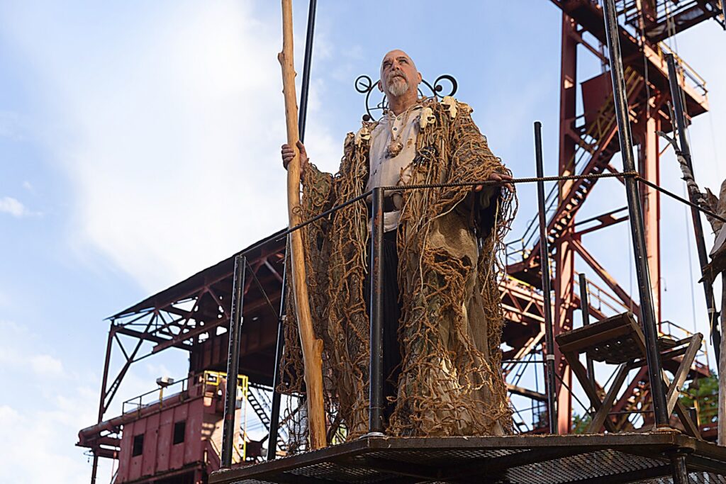 A man in a brown robe holds out a large brown branch on a red industrial set.