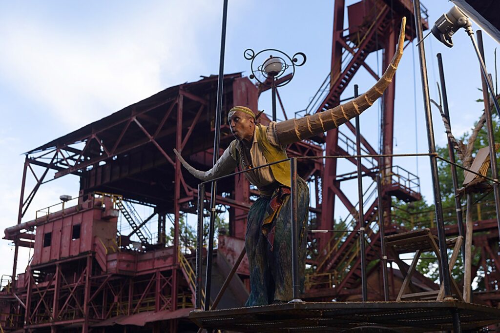 A person playing a creature with long tentacle arms on a red industrial set.