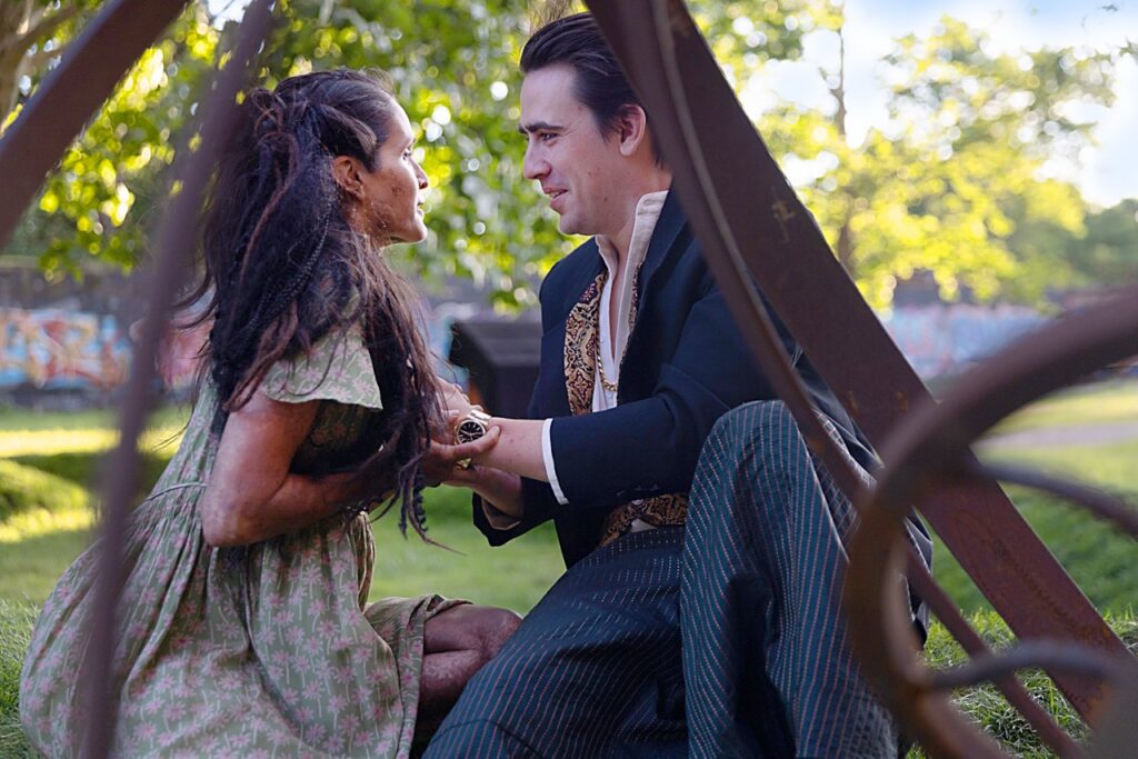 A man holds a woman's hands behind a sculpture outside as they look at each other.
