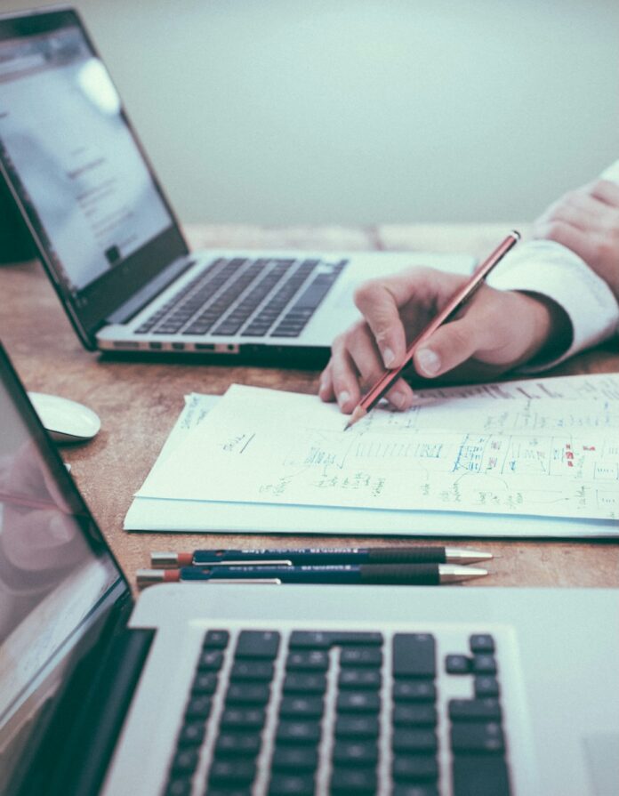 A person writing financial information on a sheet of paper between two laptops.