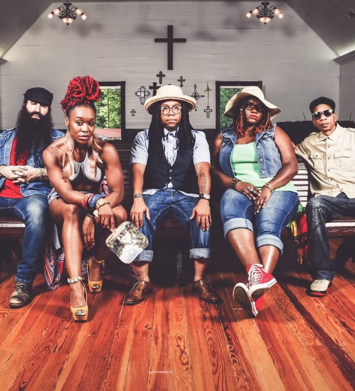The rap group members of Arrested Development sit in a church pew with a wooden floor beneath them and cross above them.