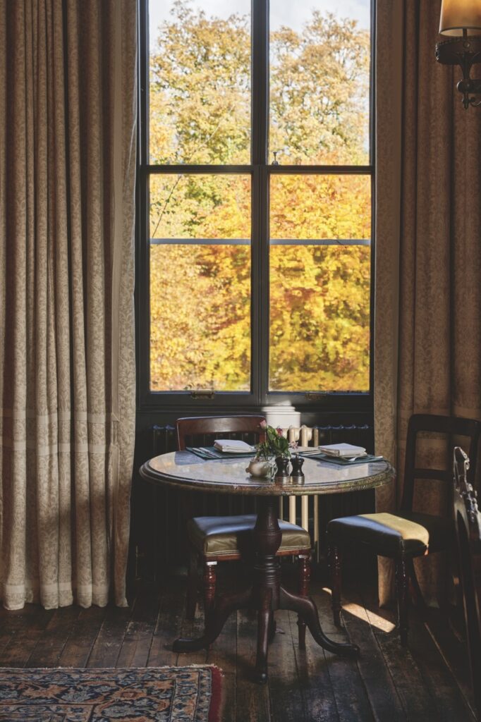 A table and chair sit by a window and long curtains.