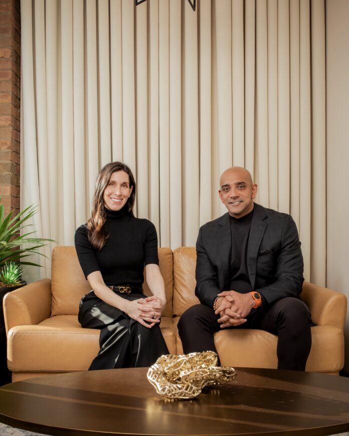 Sunjay Mannan and Sophia Moses sit on a beige couch in black clothing with a table in front of them.