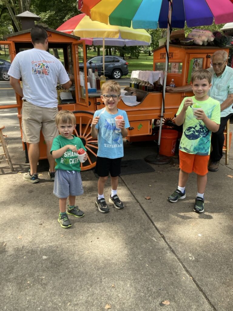Three kids stand in front of Gus and YiaYia's ice treat stand with rainbow umbrellas.