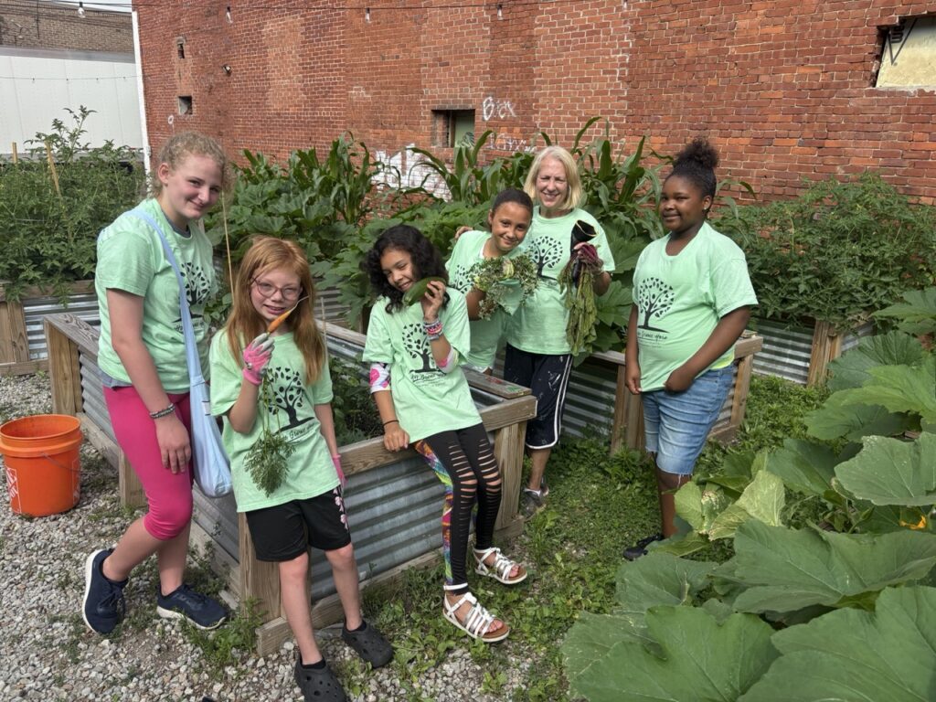A group of kids in green shirts and a woman holding vegetables in a garden.