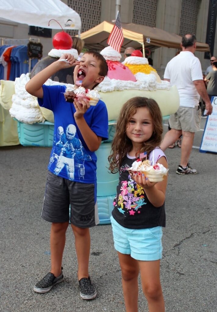 Two kids hold banana split as the boy puts the cherry into his mouth.