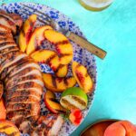 Sliced chipotle rubbed pork tenderloin, grill-marked peaches, and lime on a blue speckled plate with a vintage fork.