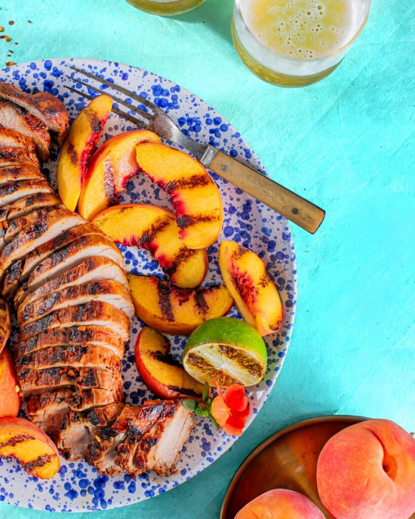 Sliced chipotle rubbed pork tenderloin, grill-marked peaches, and lime on a blue speckled plate with a vintage fork.