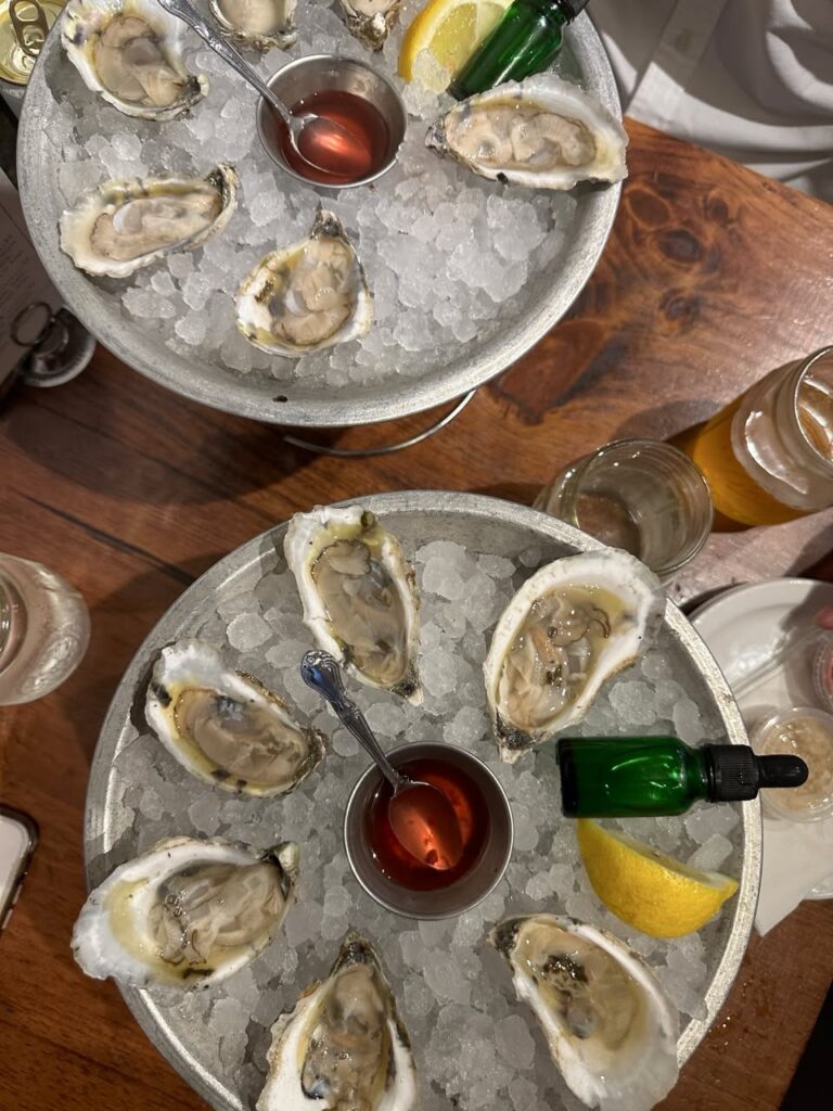 Two steel plates of oysters on ice with sauce and lemon slices.