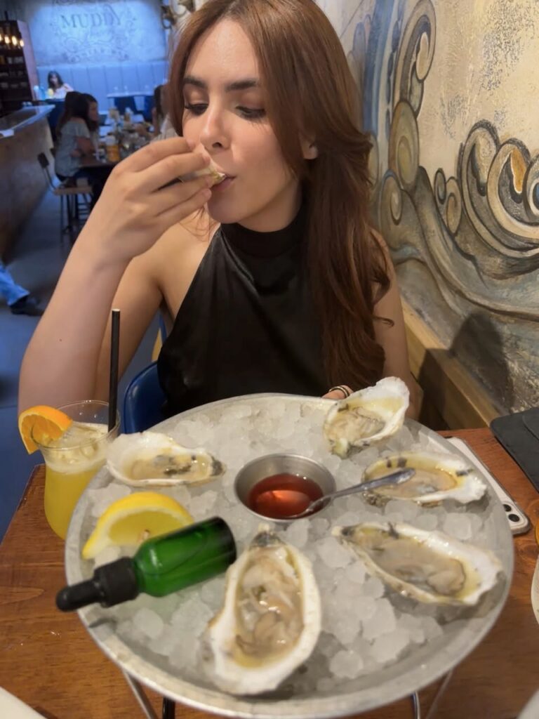 Ashley sits at a table slurping on an oyster from the tray in front of her.