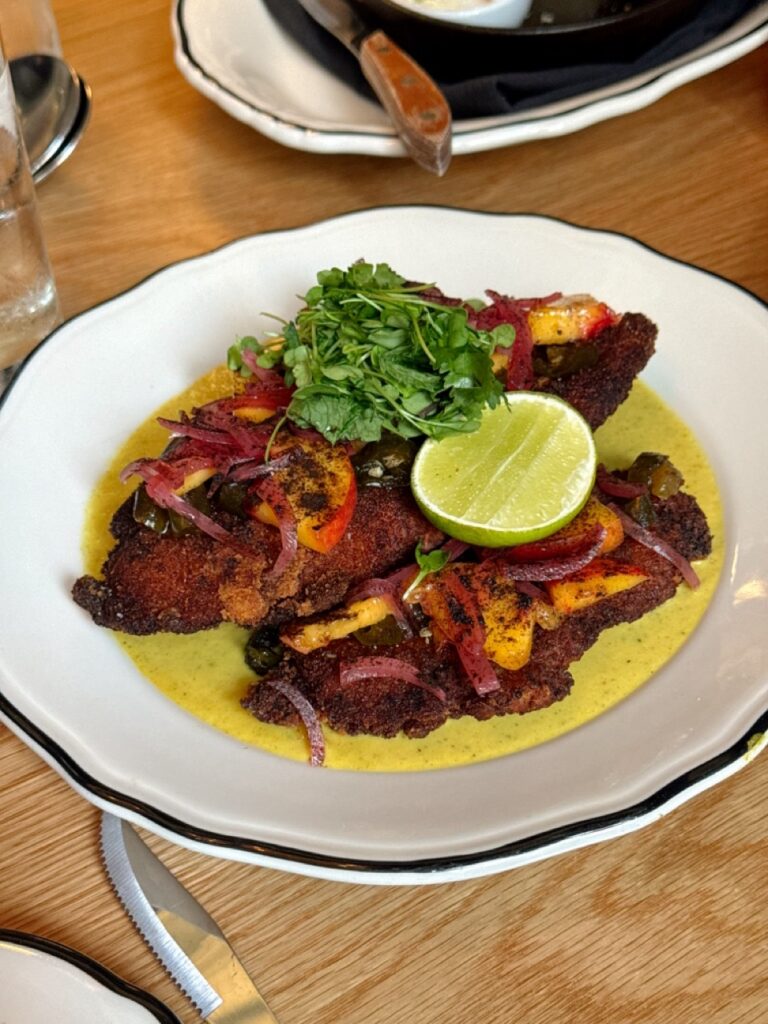A white plate on a brown table with chicken katsu and a life on top and herbs.