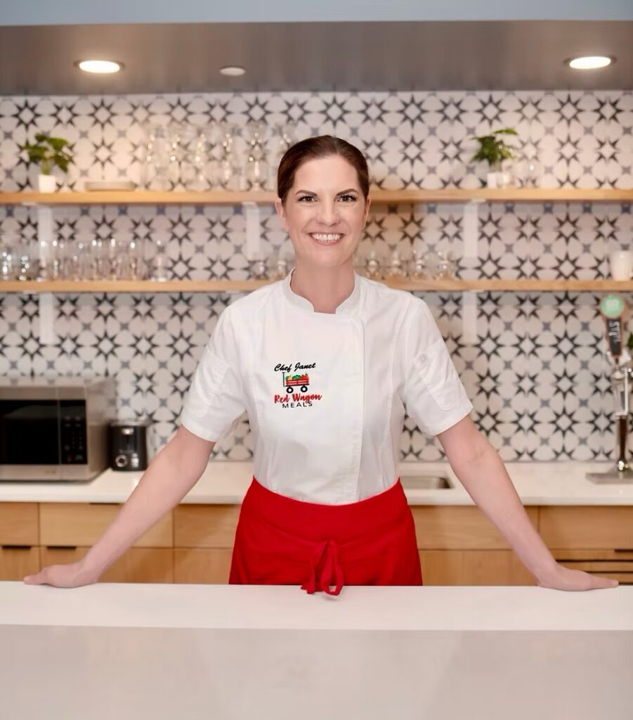 Janet stands at a counter with a white chefs shirt and red apron around her waist.
