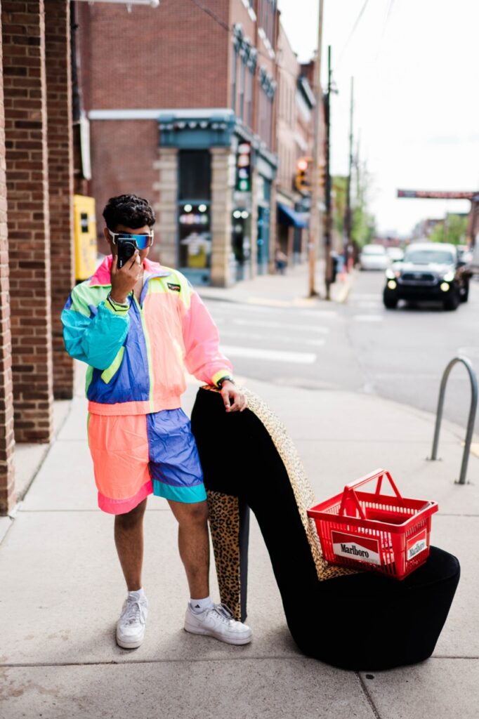 Arjun stands in a vintage color block exercise set holding up a camera beside a giant high heel and shopping basket.