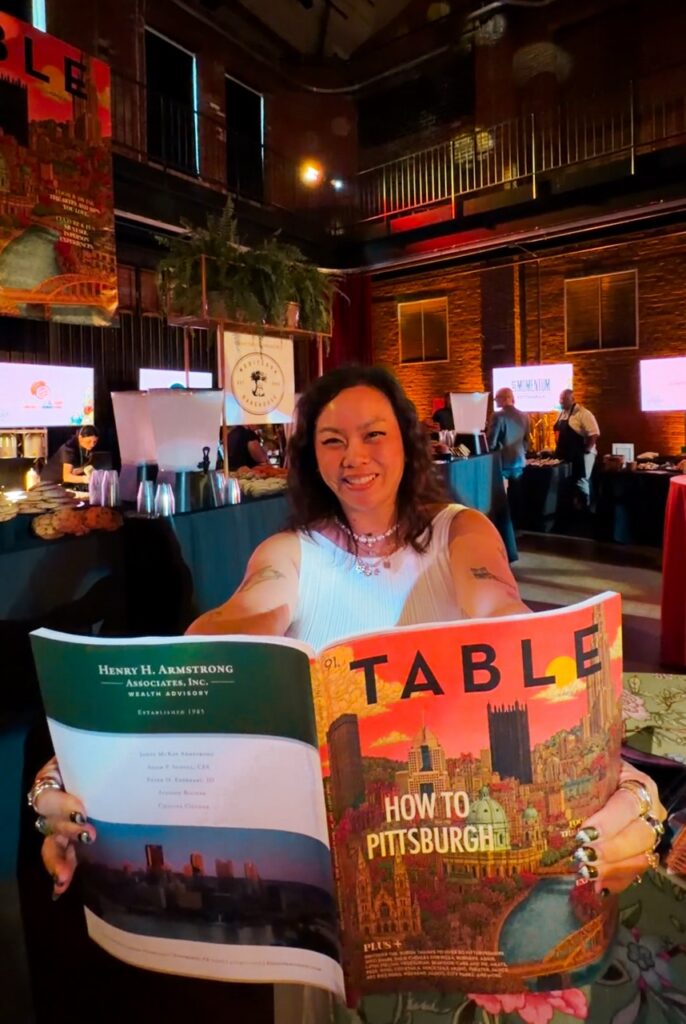 Soph sits at a table reading a table magazine.